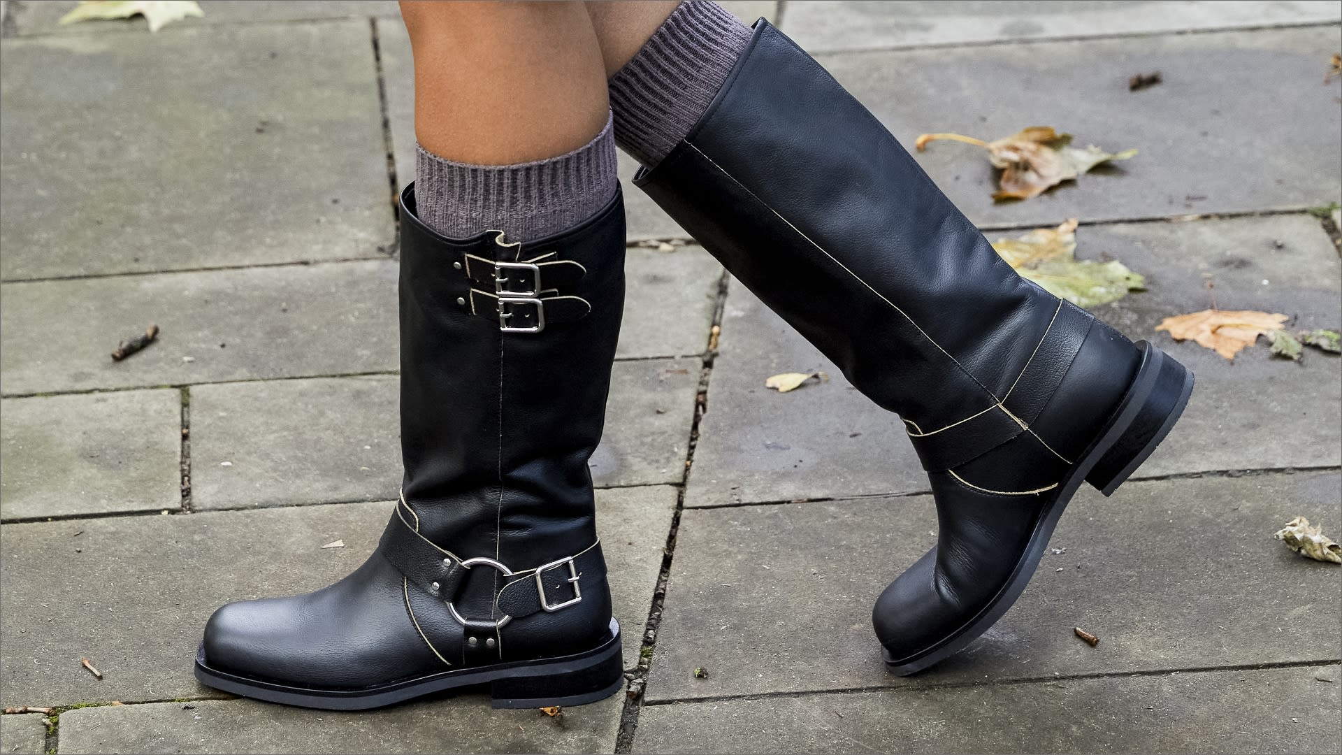 Woman wearing black flat knee high biker boots with buckle detail paired with grey knee high socks